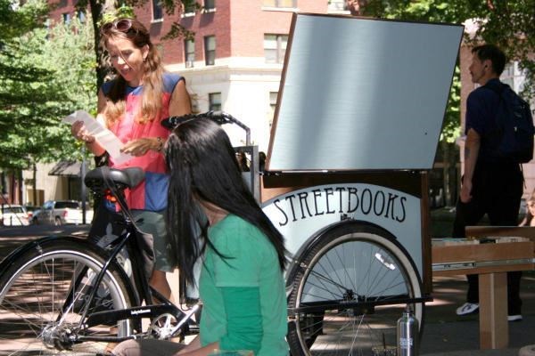 Laura Moulton (standing) pedals her bicycle-powered Street Books library to locations around Portland, Ore., providing books to the city's homeless, who don't qualify for library cards.  <P>Courtesy of Ben Parzybok